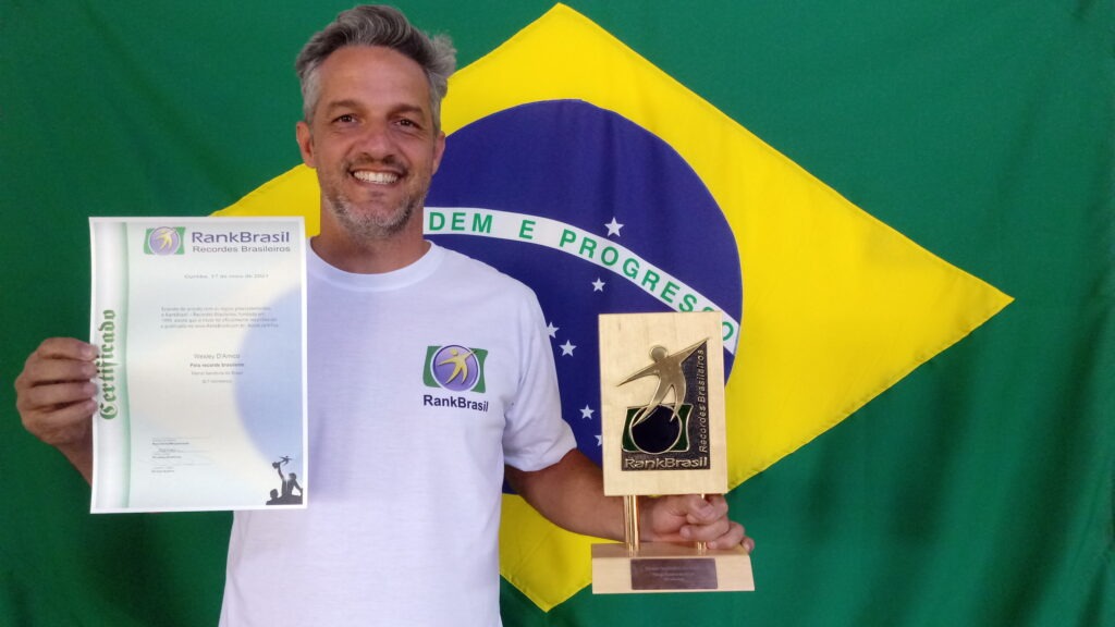 Artista Wesley D'Amico segurando seu certificado. Ao fundo podemos ver a bandeira do Brasil.